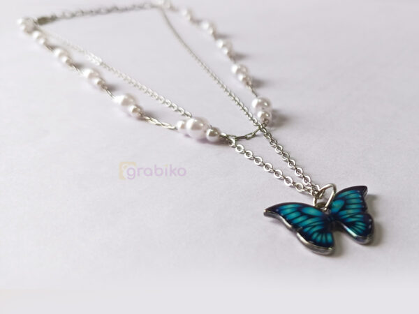 Two-layer necklace featuring a pearl choker chain and a long silver chain with a blue butterfly pendant, worn by a modern woman.